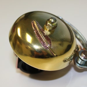 May include: A gold bicycle bell with a decorative feather design. The bell is attached to a black handlebar mount.