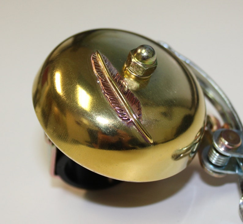 May include: A gold bicycle bell with a decorative feather design. The bell is attached to a black handlebar mount.