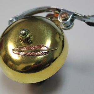 May include: A gold bicycle bell with a small feather design on the front. The bell has a silver lever and a green knob.