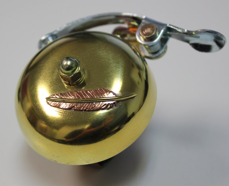 May include: A gold bicycle bell with a small feather design on the front. The bell has a silver lever and a green knob.