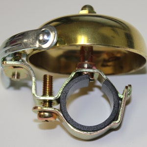 May include: A gold bicycle bell with a black rubber clamp. The bell has a chrome-plated bracket and a screw for attaching it to a handlebar.