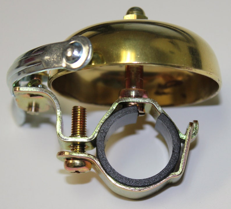 May include: A gold bicycle bell with a black rubber clamp. The bell has a chrome-plated bracket and a screw for attaching it to a handlebar.
