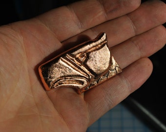 May include: A copper-colored metal pendant with a detailed design of a stylized face. The pendant is smooth and has a shiny finish.