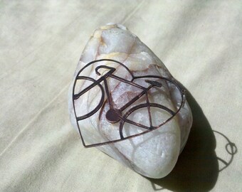 May include: A heart-shaped rock with a metal cutout of a bicycle inside. The rock is white with brown and yellow veins.