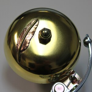 May include: A gold bicycle bell with a decorative feather design. The bell has a round shape and a silver metal bracket.