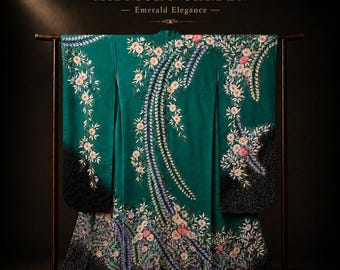 May include: Emerald green kimono with floral embroidery, displayed on a wooden rack. The kimono features a detailed design of flowers and foliage in shades of pink, white, and blue. The text "MIDNIGHT GARDEN - Emerald Elegance -" is at the top.