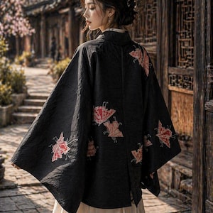 Puede incluir: Una chaqueta negra estilo kimono con bordados florales rojos y beige. La chaqueta tiene mangas anchas y se usa sobre una falda color crema. La imagen está tomada al aire libre con un fondo borroso de un edificio asiático tradicional.