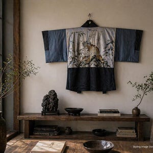 May include: A traditional Japanese kimono with a tiger and bamboo design. The kimono features a color block design with navy blue sleeves and a black bottom. The background is a light gray with a detailed illustration of a tiger and bamboo.
