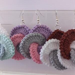 Può includere: Una collezione di orecchini all'uncinetto fatti a mano in vari colori, tra cui bianco, blu, rosa, grigio, viola e marrone. Ogni orecchino presenta un design a spirale e ganci argentati. Un accessorio unico.
