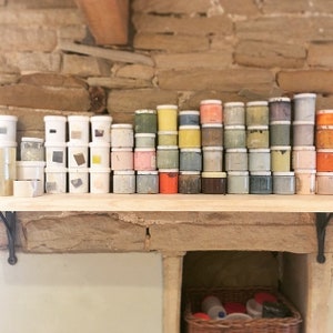 May include: A shelf filled with small jars of paint in various colors, including white, yellow, orange, red, pink, green, blue, and brown. The jars are arranged in rows and columns, creating a colorful and organized display.