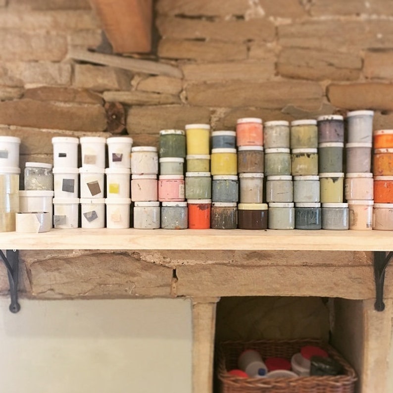 May include: A shelf filled with small jars of paint in various colors, including white, yellow, orange, red, pink, green, blue, and brown. The jars are arranged in rows and columns, creating a colorful and organized display.