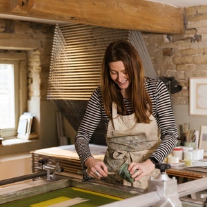 May include: A woman in a striped shirt and apron is using a squeegee to apply green ink to a screen printing frame. The frame is on a table with other art supplies.