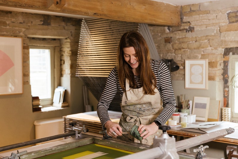 May include: A woman in a striped shirt and apron is using a squeegee to apply green ink to a screen printing frame. The frame is on a table with other art supplies.