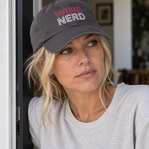Puede incluir: Una gorra de béisbol gris carbón con el texto WINE NERD bordado en la parte delantera. La palabra WINE está cosida en rojo, mientras que la palabra NERD está cosida en blanco. La gorra tiene una visera curva y una corona estructurada con botón.
