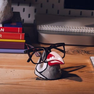 May include: A whimsical ceramic glasses holder shaped like a bird's head, painted in gray, white, and red. The holder is designed to cradle eyeglasses, with a pair of tortoiseshell glasses resting on it. The holder sits on a wooden desk.