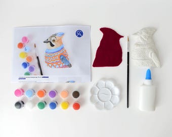May include: A craft kit with a bird theme, featuring a printed illustration, a felt cutout, a ceramic bird figurine, paint pots in various colors, a paintbrush, a palette, and glue. The illustration depicts a bird wearing a sweater.
