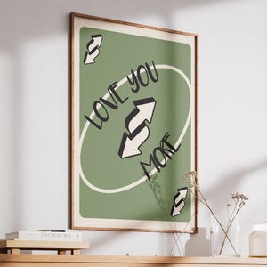 May include: A framed art print featuring a green background with the words "LOVE YOU MORE" in a handwritten style. The design includes a white oval and reverse arrow symbols. The print is displayed on a wooden shelf.