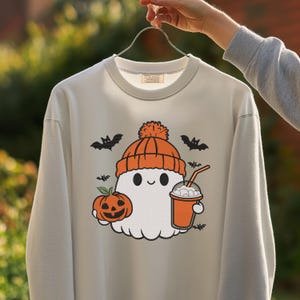 Pode incluir: Um moletom cinza claro com um design de Halloween. O design inclui um fantasma amigável usando um gorro laranja, segurando uma bebida e uma abóbora. Morcegos pretos estão espalhados ao redor do fantasma. O moletom é segurado por um cabide de metal.