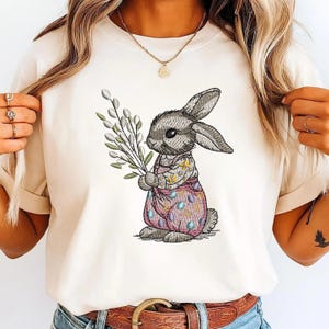 May include: A cream-coloured t-shirt with an embroidered Easter bunny holding a bundle of pussy willow branches. The bunny wears a pink and blue patterned outfit with egg designs. The design is centered on the shirt.