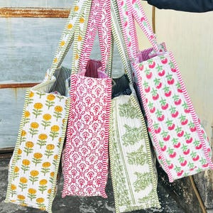 May include: A selection of patterned tote bags in various colours. The bags display floral and fruit designs on a white background. Patterns include yellow flowers, red florals, green leaves, and pink strawberries. Each bag has matching patterned straps.
