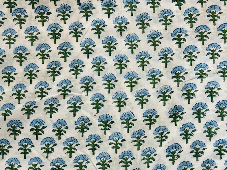 May include: White fabric with a repeating pattern of blue floral designs. Each flower has a detailed petal arrangement and is set against a backdrop of green stems and leaves. The fabric is likely cotton or linen.