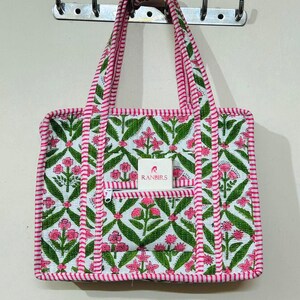 May include: A white tote bag with a floral pattern in pink and green. The bag has a zippered front pocket and pink striped trim. The handles are also pink and white striped. The brand name "RANBIRS" is visible on a small tag.