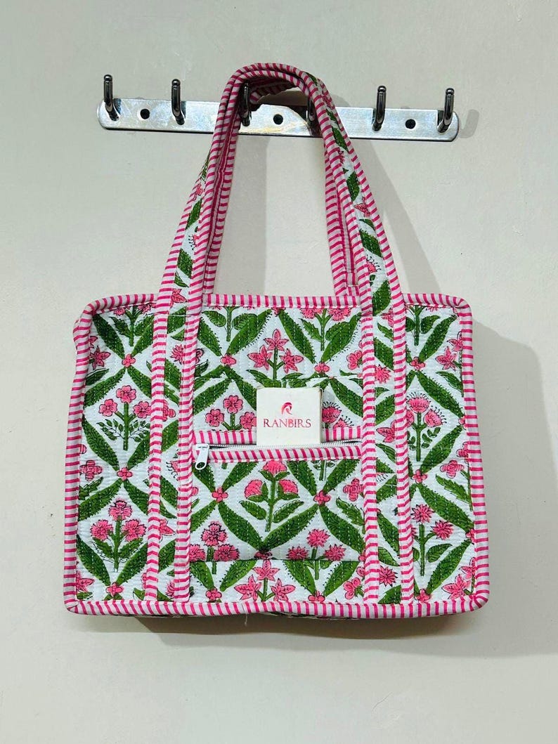 May include: A white tote bag with a floral pattern in pink and green. The bag has a zippered front pocket and pink striped trim. The handles are also pink and white striped. The brand name "RANBIRS" is visible on a small tag.