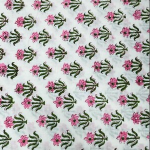 May include: White fabric with a repeating pattern of pink flowers and green leaves. The floral design is evenly spaced across the fabric, creating a consistent and visually appealing texture. The flowers have multiple petals.