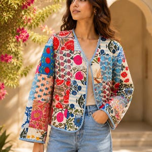May include: A quilted jacket featuring a patchwork design with various floral patterns in shades of red, blue, pink, and green. The jacket has long sleeves, an open front, and blue and white striped trim along the edges, worn with blue jeans.