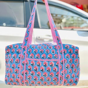 May include: A blue tote bag with a floral pattern in pink and white. The bag has pink and white striped handles and trim. The bag is held up by a person.