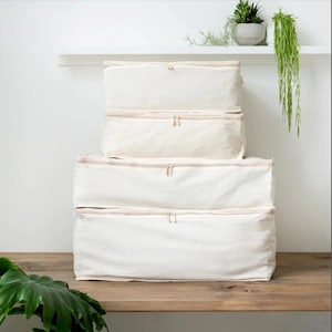 Puede incluir: Cuatro contenedores de almacenamiento de tela blanquecina apilados sobre una superficie de madera. Cada contenedor tiene un cierre de cremallera y una forma rectangular. Los contenedores son de diferentes tamaños, el más grande en la parte inferior.