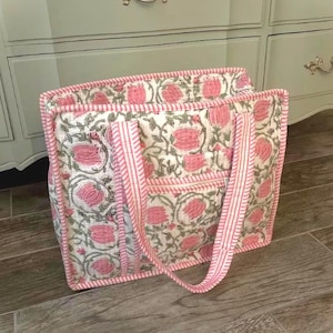May include: A quilted tote bag with a floral pattern in pink and gray on a white background. The bag has pink striped handles and trim. The bag is sitting on a wooden surface.