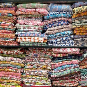 May include: Stacks of colorful fabric with floral and geometric patterns are displayed on shelves. The fabrics are in various colors, including pink, blue, yellow, and green. The image showcases a variety of textiles.