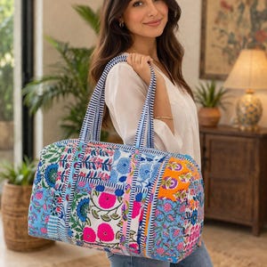May include: A colorful, patterned duffel bag with blue and white striped handles. The bag features a patchwork design with floral patterns in shades of blue, pink, and orange. The bag is being carried over the shoulder.