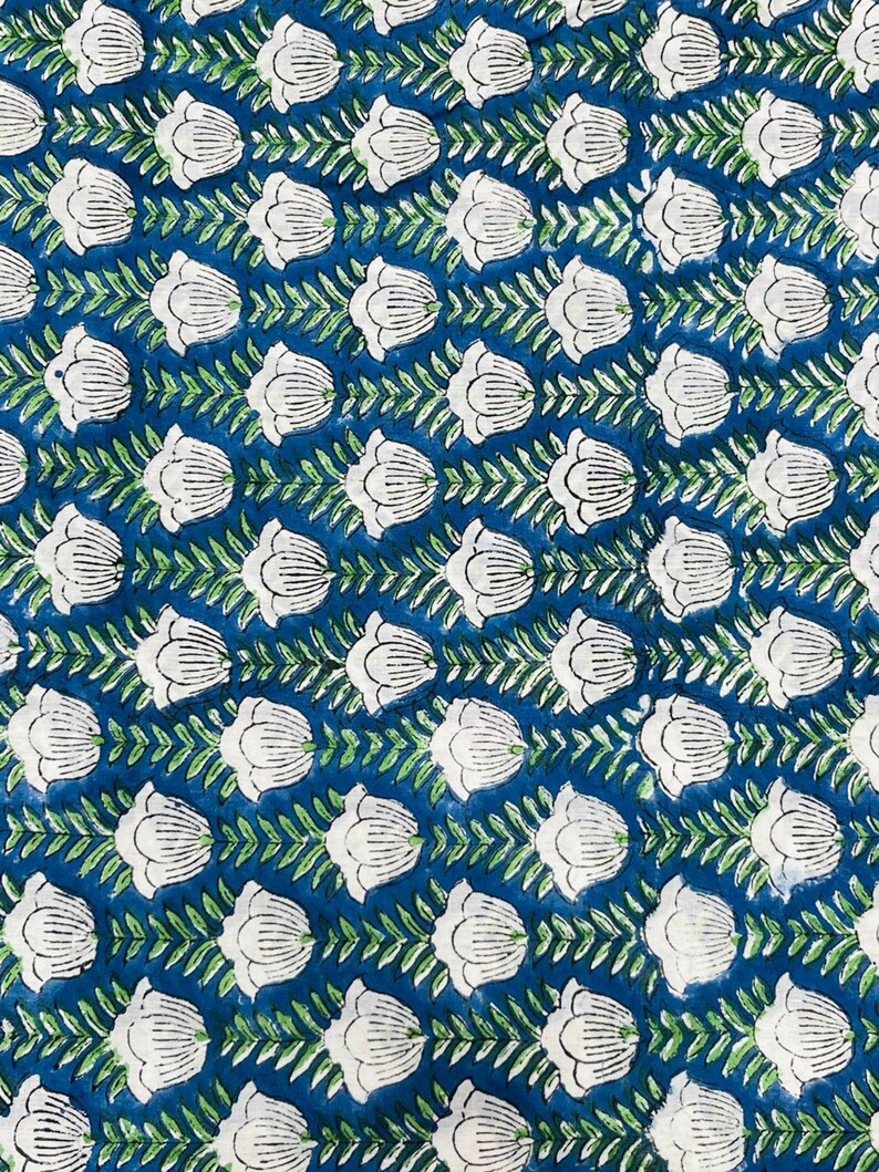 May include: A close-up of a fabric with a repeating floral pattern. White tulip-like flowers are arranged in rows against a deep blue background. Green leafy vines connect the flowers, creating a symmetrical design. The fabric has a textured appearance.