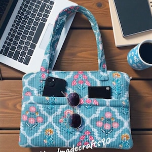 Peut inclure: Un sac fourre-tout matelassé bleu avec un motif floral rose et jaune. Le sac a deux poignées et plusieurs poches. Des lunettes de soleil, un téléphone et une carte sont visibles dans les poches. Le texte "Handmadecrafts90" est imprimé sur le sac.