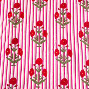 May include: A fabric textile featuring a repeating pattern of red floral motifs with green stems and leaves arranged in vertical columns. The background consists of alternating vertical stripes in bright pink and light beige, creating a vibrant contrast.