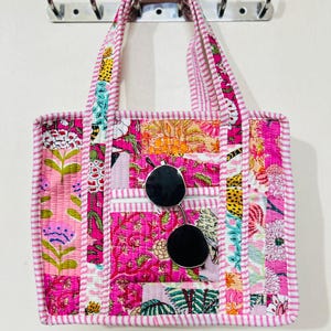 Puede incluir: Un bolso tote acolchado rosa con un diseño de patchwork floral. El bolso tiene un ribete y asas a rayas, con un bolsillo en la parte delantera. Gafas de sol negras descansan en el bolsillo. El bolso está colgado en una pared.