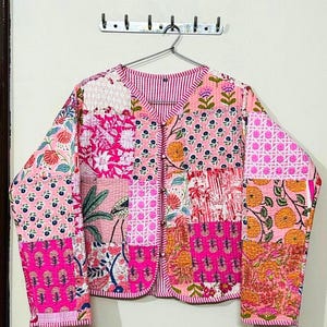 May include: A patchwork jacket in shades of pink, featuring floral and geometric patterns. The jacket has a V-neck and long sleeves. The design includes various pink hues, with accents of orange and white. The jacket is hanging on a metal hanger.