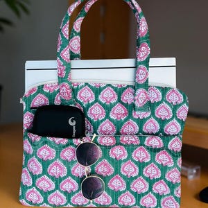 May include: A green and pink patterned tote bag with a leaf design. The bag has a zippered top, two handles, and a front pocket holding a black case and sunglasses. The bag is sitting on a wooden surface.