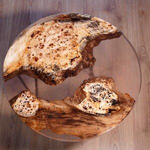 Puede incluir: Una mesa de centro redonda con una tapa de resina transparente y un diseño de madera de raíz natural. La madera presenta tonos de marrón, beige y crema, creando una estética única. La mesa está soportada por patas metálicas.