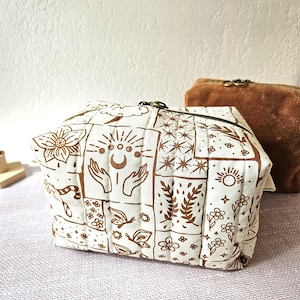 Könnte beinhalten: Eine gesteppte Kosmetiktasche in Weiß und Braun mit Reißverschluss. Die Tasche hat ein sich wiederholendes Muster aus Himmels- und Blumendesigns. Im Hintergrund ist eine zweite braune Tasche zu sehen.