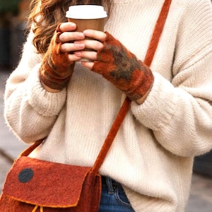 Puede incluir: Una persona con un suéter color crema, jeans azules, guantes sin dedos color óxido y un bolso cruzado a juego. El bolso tiene un detalle de círculo negro y detalles de cuerda amarilla. Sostiene una taza de café.