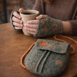 Puede incluir: Un bolso bandolera y guantes sin dedos de lana afieltrada en tonos verdes y naranjas. El bolso tiene un cierre de cordón y un botón decorativo. El conjunto está sobre una mesa de madera, evocando un estilo artesanal y acogedor.