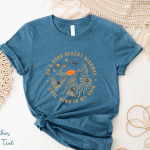 Hand-drawn graphic t-shirt featuring a dreamy desert landscape at sunset with surreal elements like planets in the sky, cacti, and animal skulls. The design includes the iconic lyric “On a Dark Desert Highway, Cool Wind in My Hair” from the classic Eagles song Hotel California. A perfect blend of retro, vintage, and boho aesthetics for music lovers and free spirits.