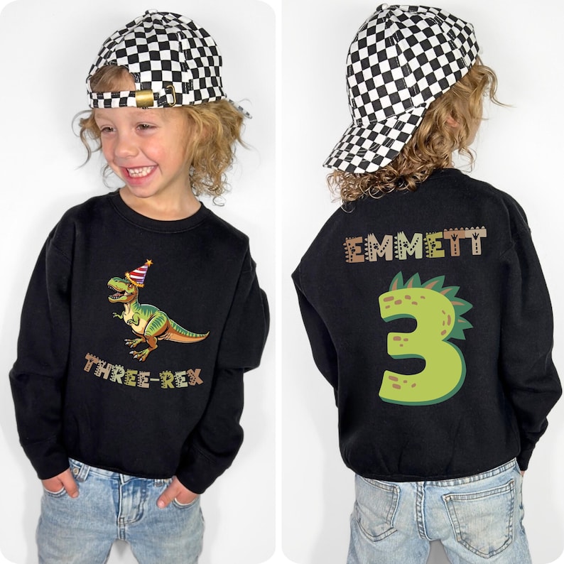 A young boy wearing a black shirt with a dinosaur and the number 3 on it, along with a checkered hat and jeans.
