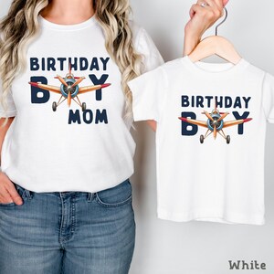 A woman wearing a white t-shirt with a graphic of a plane and the text "BIRTHDAY BOY MOM" printed on it, holding the t-shirt to the side.