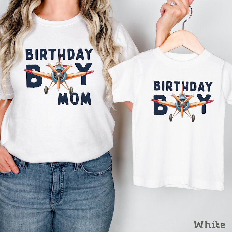 A woman wearing a white t-shirt with a graphic of a plane and the text "BIRTHDAY BOY MOM" printed on it, holding the t-shirt to the side.