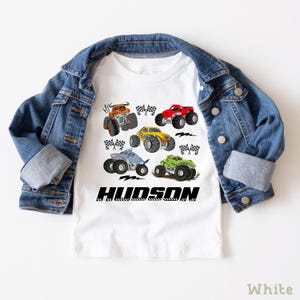 A white t-shirt with a graphic design of various monster trucks and a denim jacket are displayed on a white background.