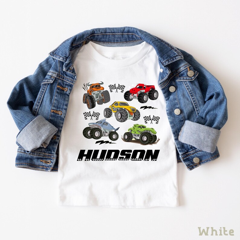 A white t-shirt with a graphic design of various monster trucks and a denim jacket are displayed on a white background.